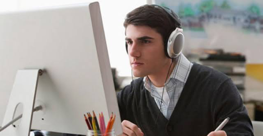 Casually Dressed Young Designer Listening To Music On Headphones While Working On A Laptop At A Desk In An Office